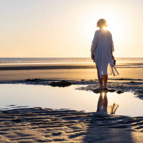 Mature woman walks along empty beach and explores tidal pools at sunset